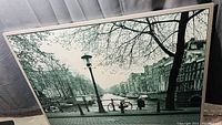 Framed black and white photo showing a canal with trees, bicycles, and buildings.