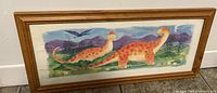 Framed kids wall decor painting featuring two dinosaurs, a pterodactyl, scenic background, and artist signature in lower left corner.