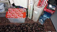 Plastic container with red Merry Christmas wrapping paper sheets and several rolls of wrapping paper standing inside and beside a clear tall container.