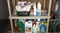 Full view of the shelf showing multiple bottles and containers of cleaning supplies with disposable cups and cloths in black basket on top shelf.