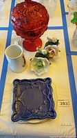 Overview of the lot items on white tablecloth showing red lidded candy dish, porcelain flowers, white mug, and blue decorative plates.