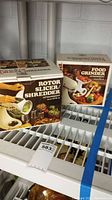 Two vintage boxed kitchen attachments: Rotor Slicer/Shredder and Food Grinder, displayed on a white metal shelf.