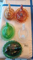Five glass hen covered dishes in various colors arranged on a white surface. Two amber/orange hens at top, one clear and two greenish hens below.