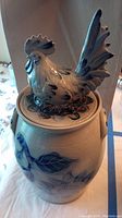 Close-up of the lidded crock showing the detailed rooster finial on top and the blue painted designs on the crock.