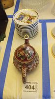 Stack of small Hummel plates with figurative designs and gold rims, next to floral decorative teapot from top view