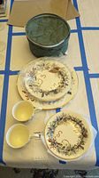 Two plates and one saucer on table with two cups and green storage container