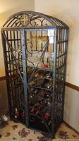 Full view of the metal wine cabinet with its ornate scrollwork doors and domed top. Shows interior with shelves and glass holders. Some wine bottles and glasses are inside but contents not included.
