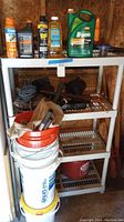 Overall view of white storage shelves holding various outside chemicals, oils, and hand tools including buckets with lids beneath the shelves.
