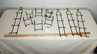 Overview of nine black metal plate holders arranged on white table including four vertical wall holders and five angled tabletop holders with measuring stick for scale