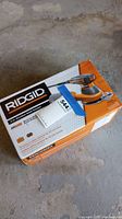 Ridgid R26011 orbital sander box on concrete floor, packaging showing product image and branding.