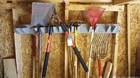 Wide shot of five garden tools hanging on wall-mounted rack, including shovels, rakes, and edger.