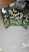 Three-panel stained glass fireplace screen showing full front view with floral and butterfly designs in green leafy background.