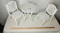 Photo showing full set of white wrought iron doll furniture including two chairs and round table on a white surface against a wall, with ruler for scale