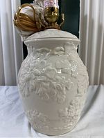 Front view of white glossy porcelain lamp base with oak leaf embossed design and attached brass lamp socket with cord wrapped up on top