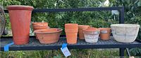 Overview of 9 terra-cotta pots of varying sizes and shapes displayed on a black metal shelf outdoors with greenery in the background.
