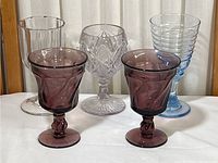 Five colorful glassware pieces including two purple goblets with twisted stems, one clear cut crystal goblet, one clear ribbed glass, and one blue ribbed glass.