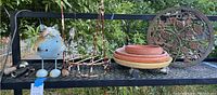 Wide shot of garden accessories collection on black metal shelf showing decorative cat figurine, metal trellis, terra cotta pots, and cast iron trivet.