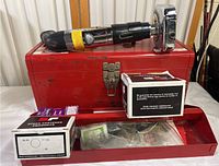 Redhead powder actuated tool resting on top of a red metal carrying case. Various boxes of small parts and accessories are nearby, some visible in the case drawer.