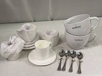 Set of four espresso cups and saucers and four spoons with two large coffee mugs stacked, all white ceramic and new with protective wrapping on cups.