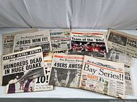 Photo showing a spread of about 15 San Francisco newspapers with big bold headlines covering the 49ers' Super Bowl win, earthquake casualties, and Giants sports news on a white surface.