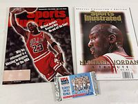 Two Sports Illustrated magazines and two connected packs of SkyBox trading cards. One magazine shows Michael Jordan dunking in a Chicago Bulls uniform (Jan 25, 1999). The other is a Special Collector's Edition titled 'Michael Jordan: A Tribute'. The trading card packs are unopened, connected side by side, showing a USA basketball team photo on the packet front.