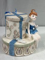 Front angled view of the porcelain ballerina music box showing the ballerina seated on the gift box base with matching blue ribbon and floral embossed design