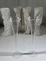 Two unwrapped giant champagne flutes with elegant hollow stems and flared rims, standing upright on a flat surface with four flutes wrapped in paper in the background.