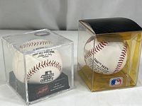 Two sealed MLB baseballs in clear protective cases side-by-side on a flat surface. The left case is clear with black base labeled 2007 All-Star Game. The right case is yellow-tinted with a black top and MLB logo at front, containing Nolan Ryan commemorative ball.