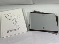 Two hardcover Rolls-Royce books lying on a white surface, one white with a line drawing of the Spirit of Ecstasy, the other gray with slipcover.