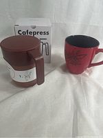 Photo showing brown Cafepress coffee press with lid and handle next to red 2007 Starbucks mug and Cafepress box in background