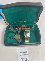 Photo showing a men's jewelry lot inside a green fabric-lined presentation case, containing a vintage gold-tone wristwatch, three pairs of cufflinks, two decorative tie pins, and a metal keychain accessory.