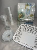 Collection of dish and decor items including square floral plates, white ceramic candlesticks, woven basket, small bowl, and metal nutcracker.