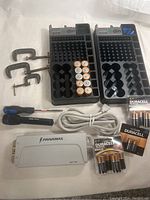 Overview shot showing two black DIN battery storage boxes partially filled with Duracell C and D batteries, two metal C-clamps, two screwdrivers, and a white Panamax power adapter with power cable.