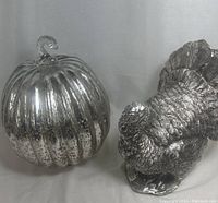 Photo showing reflective silver glass pumpkin and silver plastic turkey side by side on a white surface.