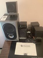 Complete set showing subwoofer, five satellite speakers, cables, and manual on table.