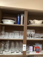 Shelf with various glassware including crystal wine glasses, short and tall drinking glasses, and some branded glasses, plus white gravy boats and plastic serveware bowls.