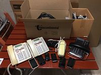 Lot on a table showing multiple landline phones, cell phones, and camera in boxes behind