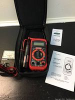 Photo showing the digital multimeter in a black case with test leads and voltage detector, with lot tag and manual visible.