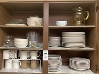 Two-shelf cabinet showing stacks of white plates, bowls, mugs and a glass pitcher