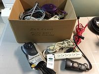 Overview photo of box with extension cords, power bars, power adapters, including one white power bar and a black mechanical timer switch on table.