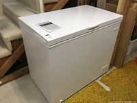 White Frigidaire chest freezer, rectangular, placed on a basement floor near stairs and yellow carpet. Front view showing brand logo on right side and power controls at bottom right.