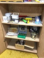 Overall view inside wood cabinet showing multiple household items arranged on three shelves including mugs, an electric hot pot, cleaning brushes, boxes and miscellaneous household goods.