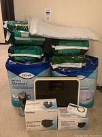 Photo showing stacked packs of Tena ProSkin Stretch and Depend incontinence briefs with a dial weigh scale and MedSup Canada box of disposable masks and pulse oximeter.