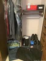 Closet with several men's dress jackets and dress shirts hanging in plastic garment bags, jeans and slacks folded on lower shelf, multiple pairs of black shoes and slippers size 12 on the floor