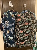 Two men's Hawaiian shirts hanging on hooks, showing colorful tropical patterns with vehicles and palm trees