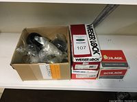 Top view of various lock parts in a cardboard box next to three boxed locksets, two Weiser Lock and one Schlage lockset.