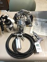Overview of two spray paint applicator guns with attached metal paint containers and hoses, plus instruction manual on table.