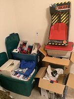 Photo showing plastic storage bins and cardboard boxes containing various Christmas decorations and loose items, plus unopened Oskar snow shovel box