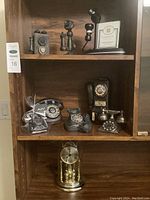 Shelf with decorative vintage style phone replicas including rotary dial models, multiple finishes, and a black and brass mantle clock under glass dome.