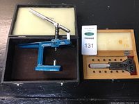 Set of watchmaker's press and watch back case opener in their respective boxes. Press is blue metal with white handle. Opener boxed in wood with foam lining and multiple bits.